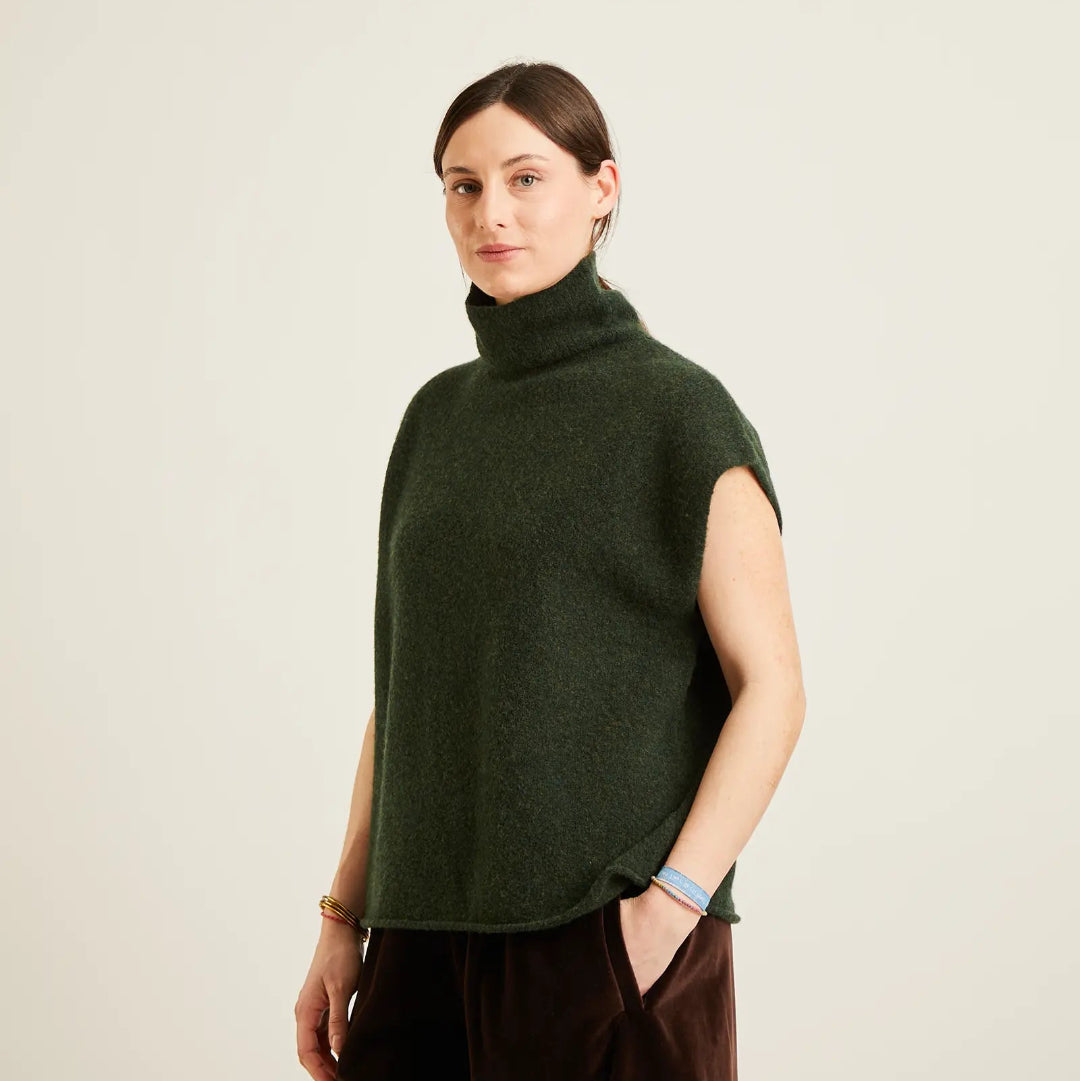 A woman wearing a dark forest green sleeveless mock neck sweater and dark brown trousers, standing against a plain light beige studio background.