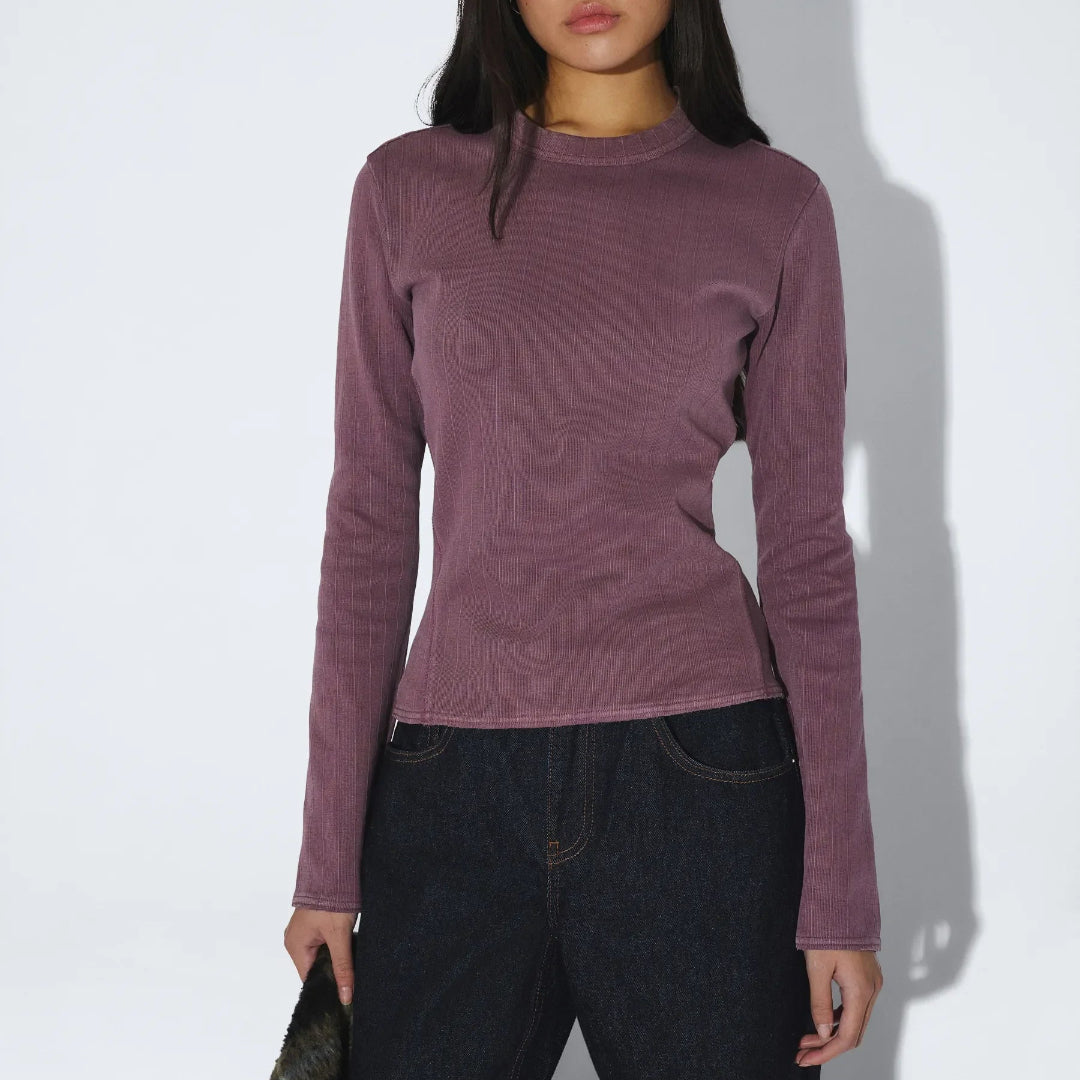 A woman modeling a fitted, long-sleeve ribbed knit top in a muted mauve-purple color, paired with dark blue jeans, against a plain white studio background with a soft shadow.