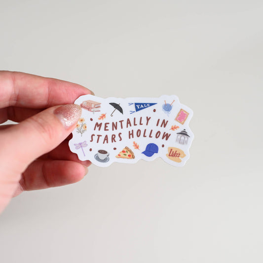 Hand holding a sticker with 'Mentally in Stars Hollow' text and graphics on a plain background
