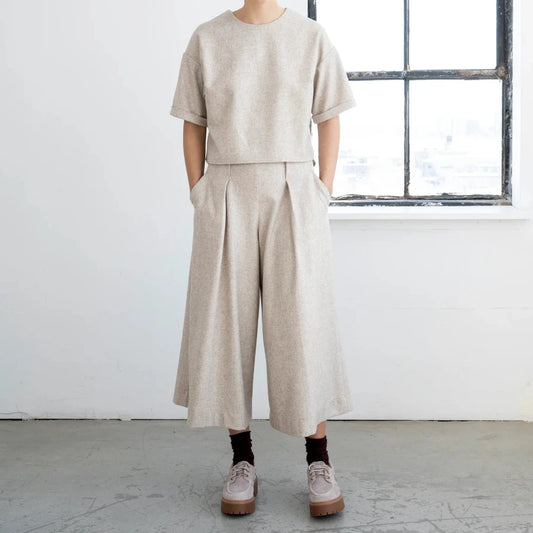 A model stands in a bright, minimalist room wearing a light beige two-piece set consisting of a short-sleeve crewneck top and matching wide-leg cropped trousers. The model is also wearing black socks and beige sneakers.