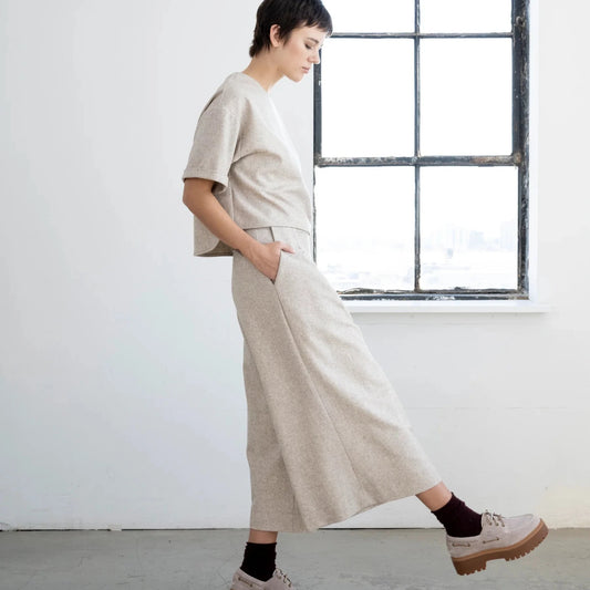 A model wearing a beige short-sleeve t-shirt and matching wide-leg cropped trousers, paired with beige platform shoes and black socks, standing in a bright room with a large window.