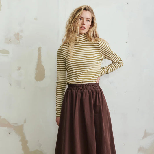 A woman stands against a textured off-white wall wearing a olive green and white striped mock-neck long-sleeve top and a dark brown pleated maxi skirt, paired with dark brown slip-on shoes.