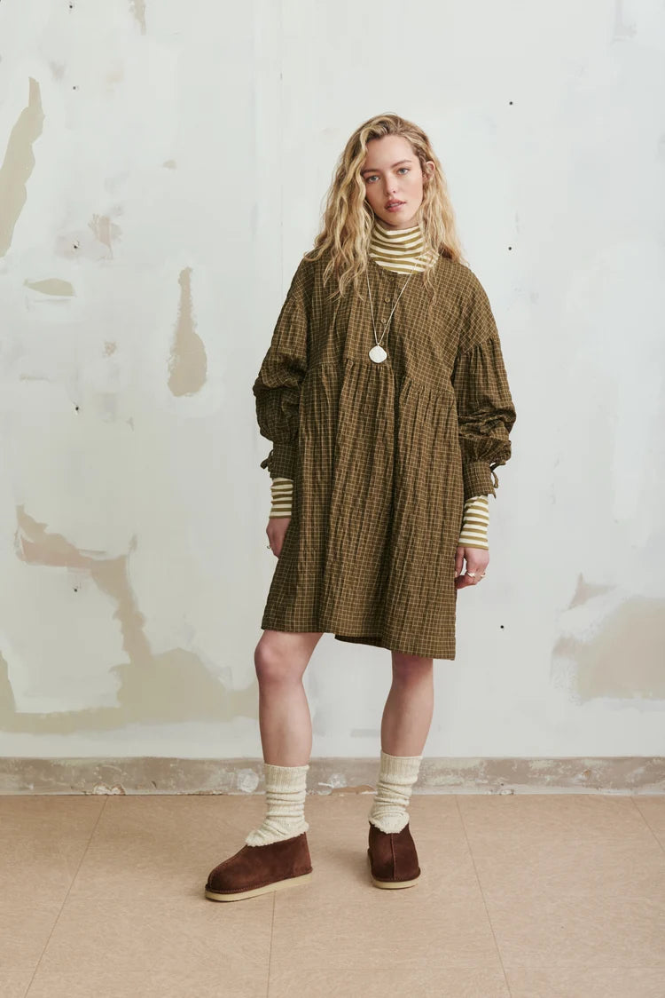 A woman stands against a textured off-white wall wearing a brown and olive checkered tunic dress over a cream striped turtleneck, cream socks, and brown suede slip-on shoes.