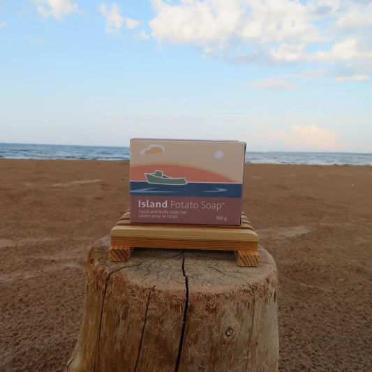 A boxed bar of Island Potato Soap stands on a small wooden platform atop a tree stump, with a sandy beach and ocean in the background under a blue sky.
