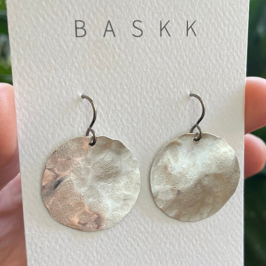 A hand holding a white BASKK jewelry display card featuring a pair of hammered round silver-tone drop earrings, with green potted plants in the background.
