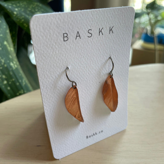 A pair of copper drop earrings, displayed on a white BASKK brand card stand on a light wooden table with a blurred green plant in the background.