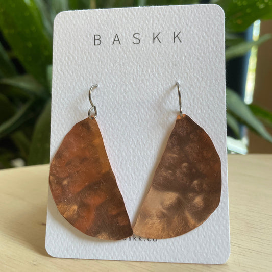 A pair of hammered copper drop earrings in a half circle design displayed on a white BASKK brand card, set against a blurred background of green foliage and a light wood surface.