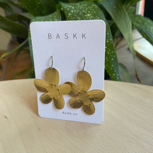 A pair of gold flower-shaped dangle earrings displayed on a white BASKK brand card, resting on a light wood surface with green houseplants in the background.