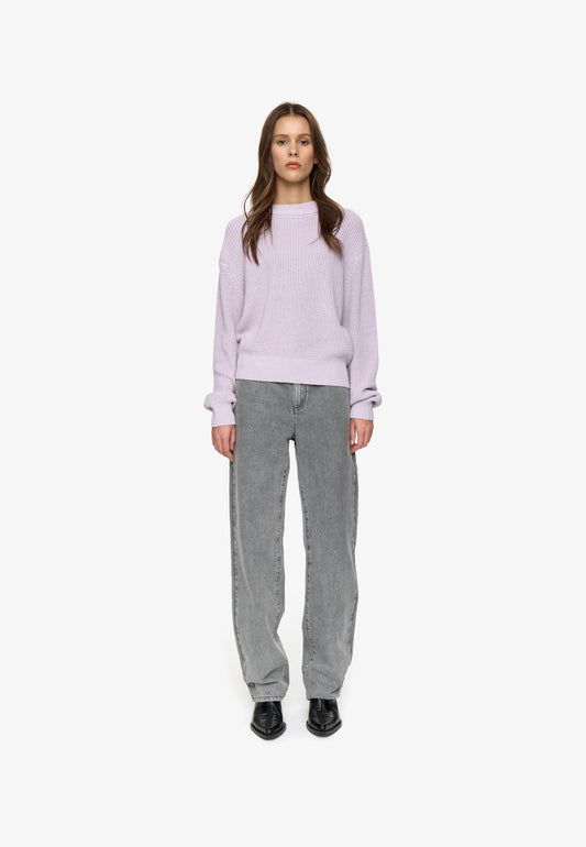 A woman stands against a plain white studio background wearing a lilac crewneck sweatshirt, gray wide-leg jeans, and black shoes.