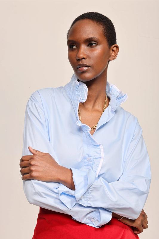 A model wearing a light blue long-sleeve button-up shirt with a ruffled collar and cuffs, paired with a bright red skirt, against a solid beige background.