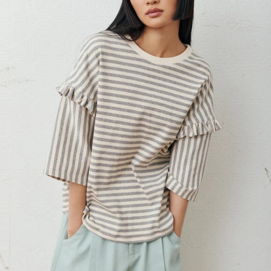 A woman with a straight black hair wearing a cream and gray striped ruffle-sleeve top and light blue trousers, standing against a white textured wall.