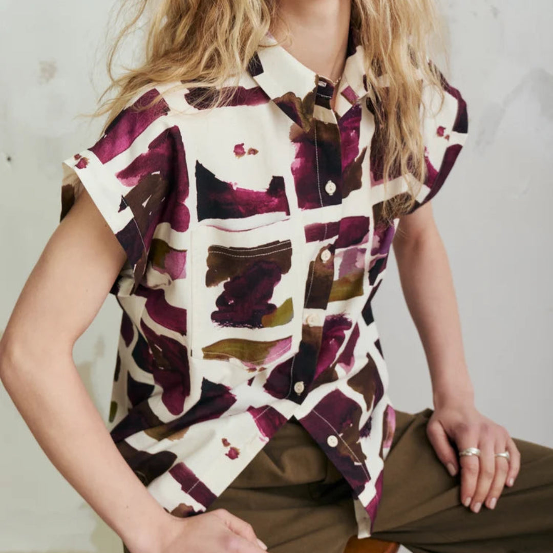 A woman wearing a short-sleeve button-up shirt with an abstract watercolor pattern in deep purple, brown, and olive green on an off-white base, paired with olive green trousers, seated against a textured light gray wall.