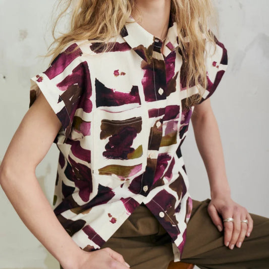 A woman wearing a short-sleeve button-up shirt with an abstract watercolor pattern in deep purple, brown, and olive green on an off-white base, paired with olive green trousers, seated against a textured light gray wall.