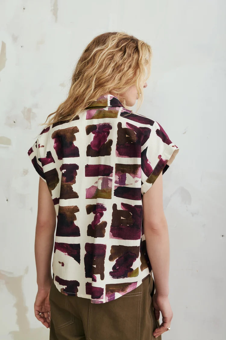 A woman viewed from behind wearing a short-sleeve button-up shirt with an abstract block pattern in deep purple, burgundy, and olive green on an off-white background, paired with brown pants against a textured white wall.