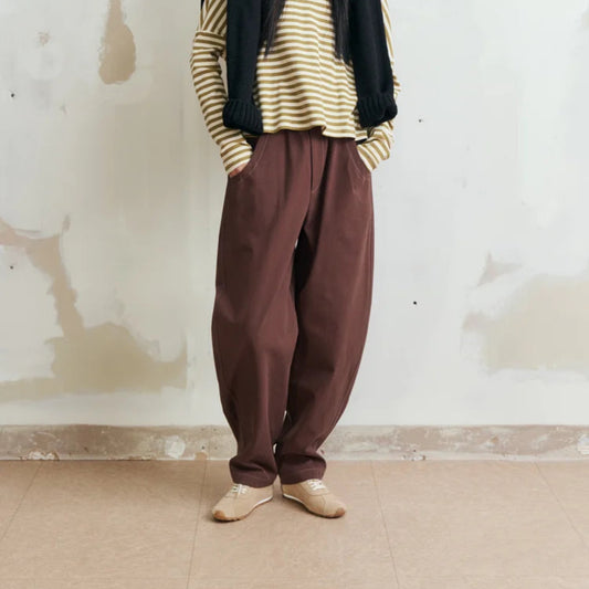 A model stands against a textured off-white wall wearing a striped beige and green long-sleeve top, a black sleeveless vest, loose brown pants, and beige shoes.