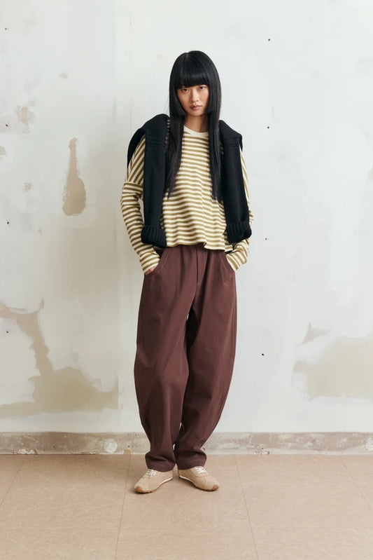 A model stands against a textured off-white wall wearing a striped beige and green long-sleeve top, a black sweater draped over the shoulders, loose brown pants, and beige shoes.