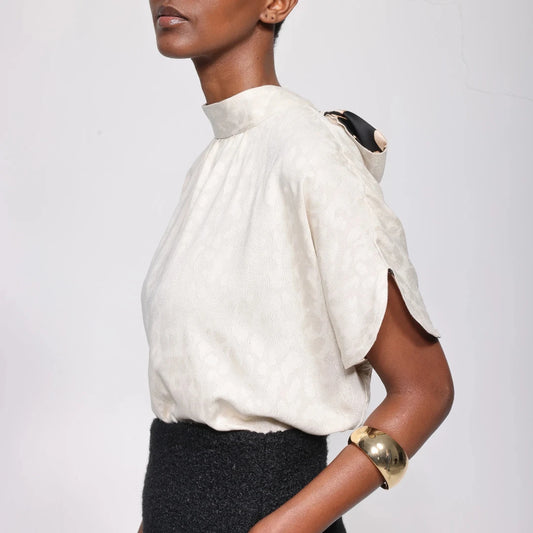 A model shown from the side wearing a textured off-white high-neck blouse with a unique shoulder detail and a black textured skirt, set against a plain light gray background.