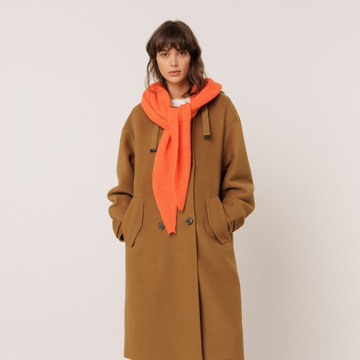 A model stands against a plain light beige studio background wearing a long camel-brown wool coat, a bright orange knit scarf, wide-leg blue jeans, and black shoes.