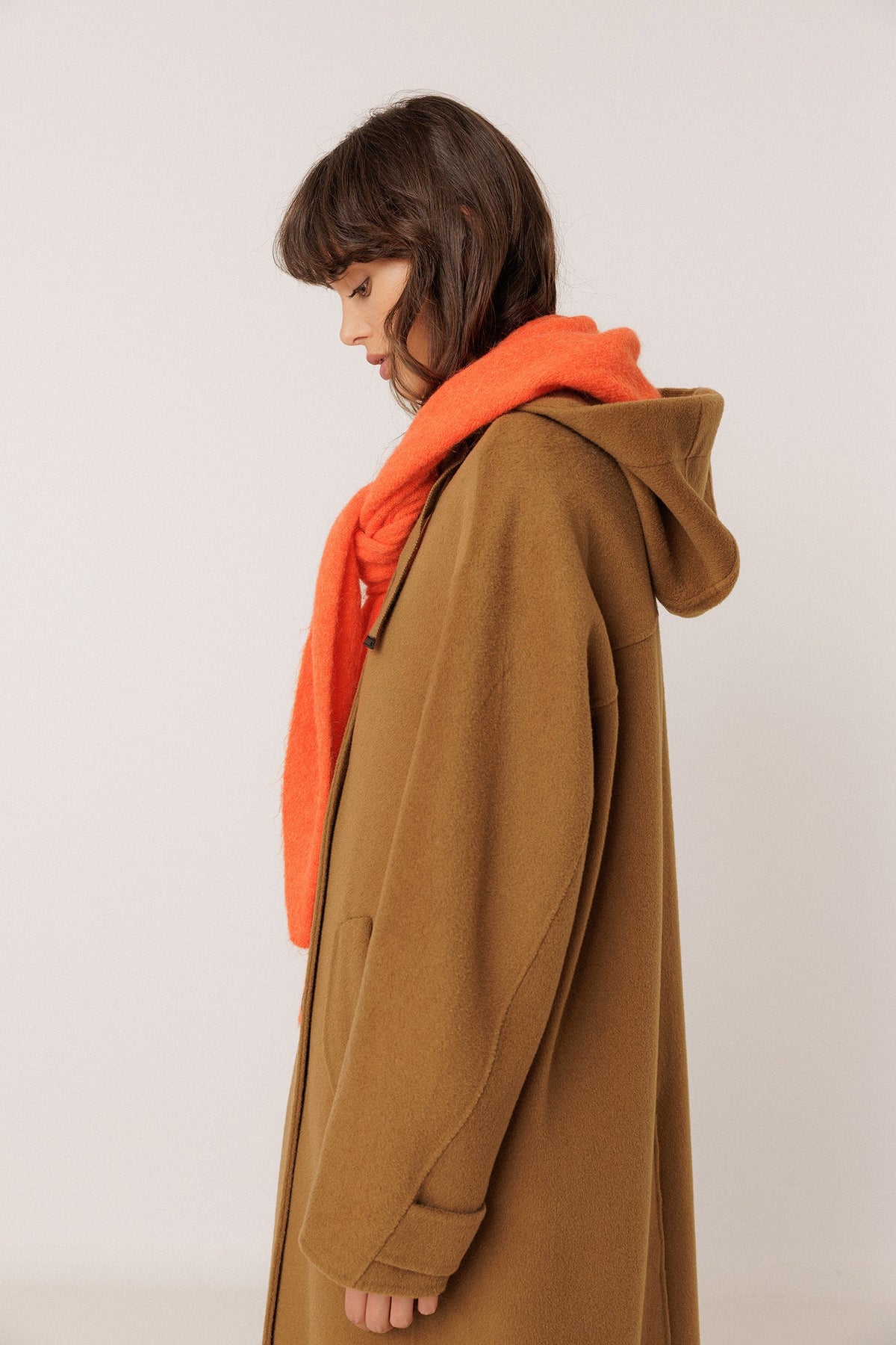 A model wearing a brown hooded coat with an orange shawl, shown from a side profile against a plain light background.