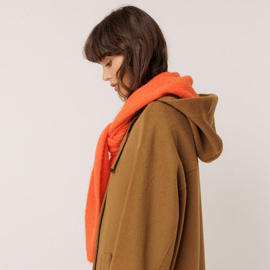 A model wearing a brown hooded coat with an orange shawl, shown from a side profile against a plain light background.