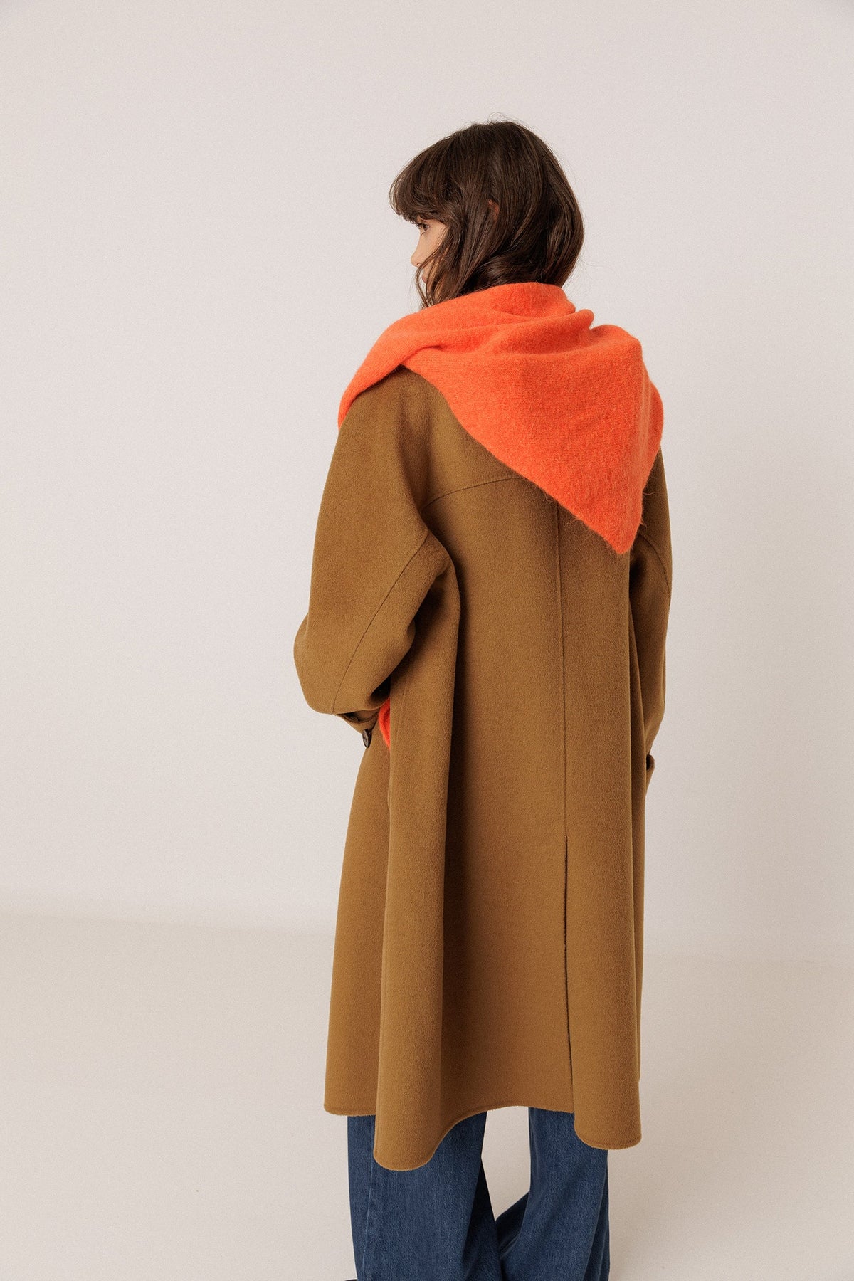 A model shown from the back wearing a camel brown coat with a bright orange shawl, paired with blue jeans, against a plain light gray studio background.