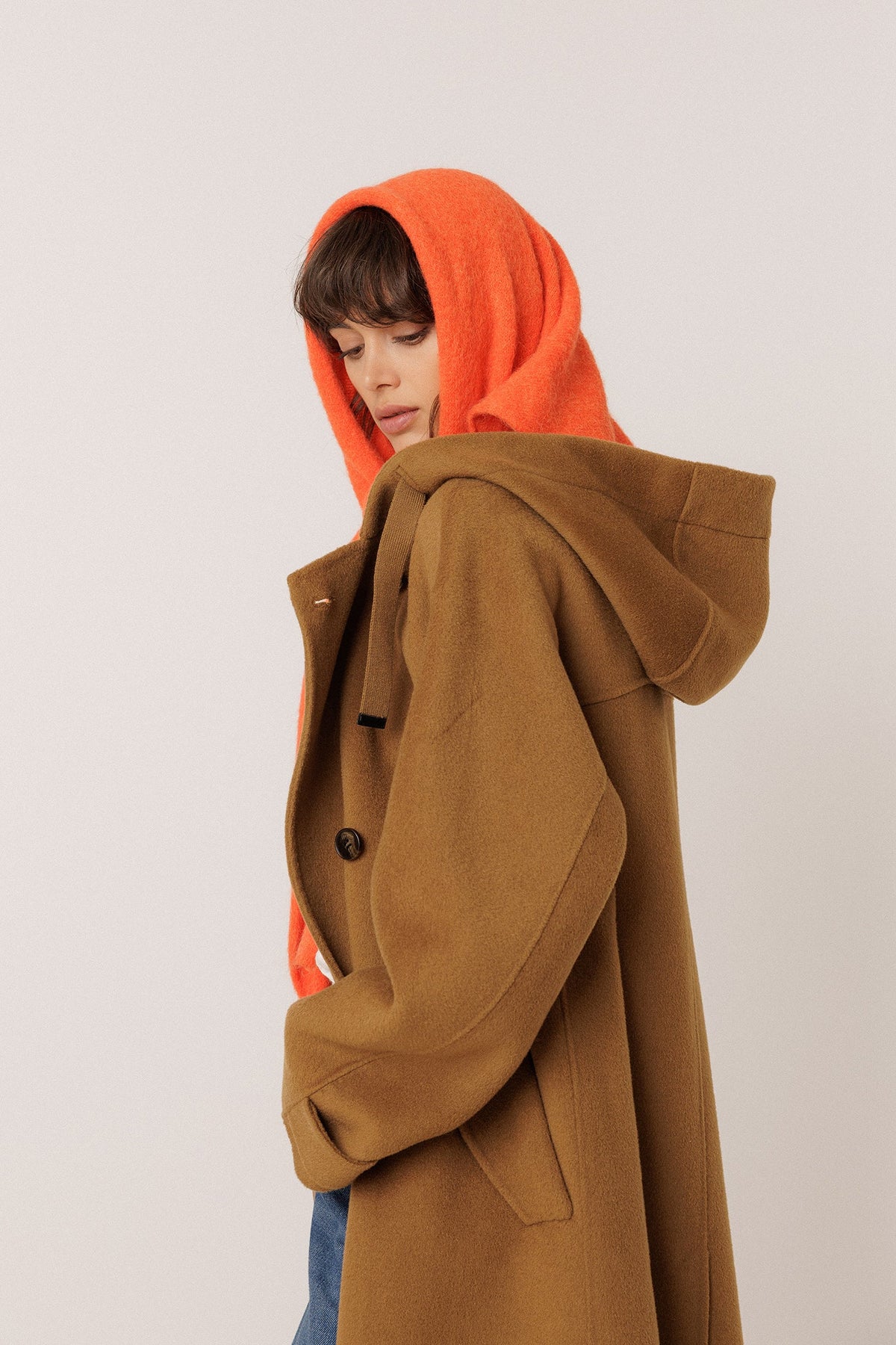 A model wearing a camel brown hooded coat with an orange shawl draped over the model's head, shown from the side against a plain light gray studio background.