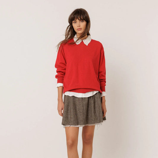 A model stands against a plain white background wearing a bright red crewneck sweater over a white collared shirt, paired with a brown textured mini skirt.
